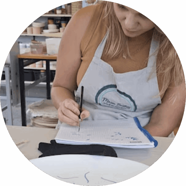 Mulher loira de avental inclinada sobre uma mesa de ateliê, usando um pincel para criar desenhos delicados em um caderno. Ao fundo, prateleiras com peças de cerâmica reforçam o ambiente de workshop criativo da Alecrim, onde arte e gastronomia se encontram para criar experiências sensoriais únicas.