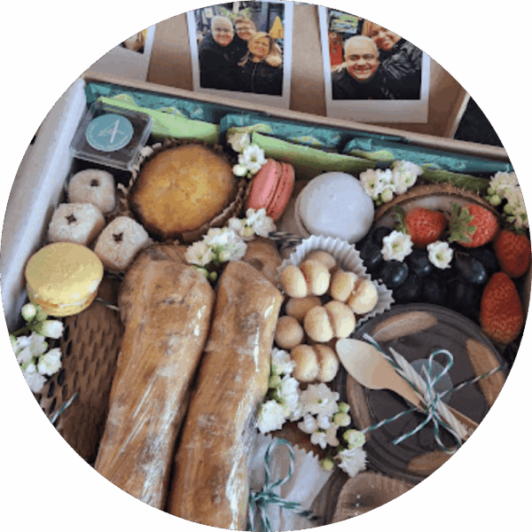 Caixa de presente artesanal da Alecrim aberta, revelando pães gourmet, macarons, frutas frescas com flores decorativas e itens de confeitaria fina. Na parte interna da tampa, fotos personalizadas estilo polaroid conectam a gastronomia ao afeto, ilustrando uma experiência de presente personalizada em Belo Horizonte.