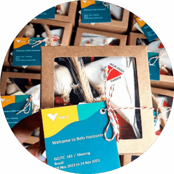 Várias caixas de presentes corporativos da Alecrim alinhadas, com foco em uma unidade frontal. A embalagem kraft possui visor transparente, revelando produtos com temática mineira e decoração rústica. Uma tag azul e amarela personalizada para a empresa Vale dá as boas-vindas a Belo Horizonte, exemplificando soluções de brindes gourmet para eventos.