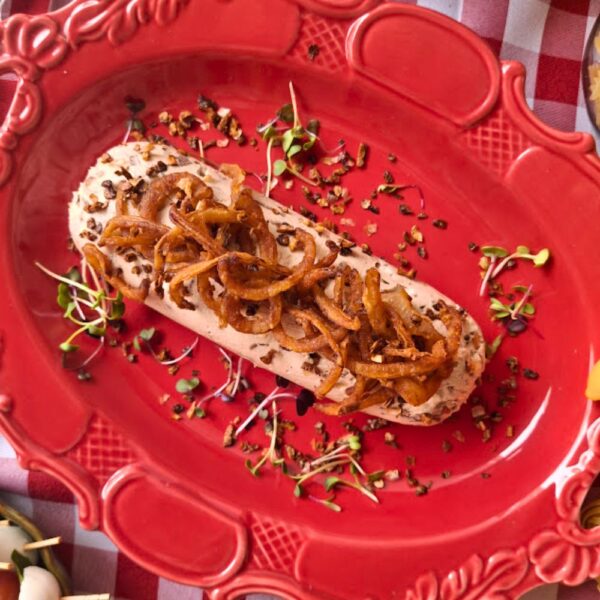 Antepasto gourmet melba de cebolas da Alecrim decorado com cebolas crocantes e brotos, servido em um prato de cerâmica vermelha ornamentada sobre toalha xadrez. A imagem destaca a gastronomia autoral e a estética artesanal para eventos e recepções intimistas em Belo Horizonte.