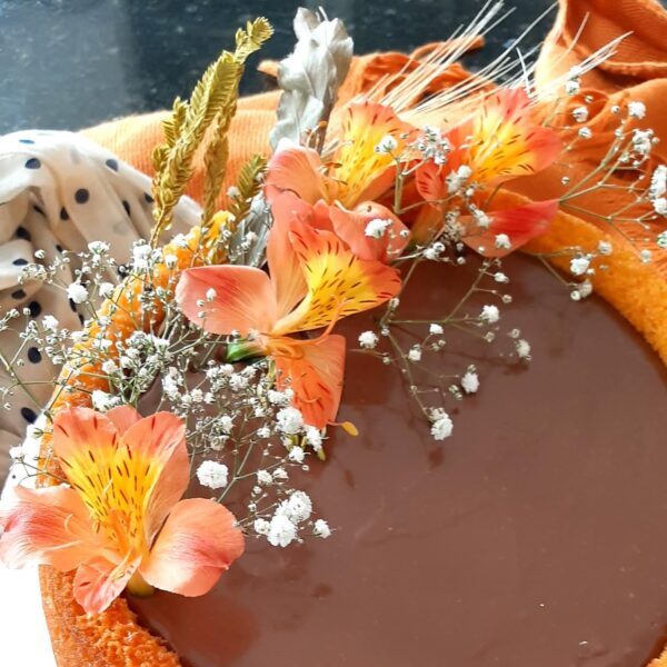 Bolo festivo de cenoura da Alecrim com cobertura brilhante de brigadeiro de chocolate. O bolo artesanal é decorado com um arranjo de flores naturais (alstroemérias) e mosquitinho, unindo a tradição do bolo caseiro com uma apresentação de luxo para eventos e cafés especiais em Belo Horizonte.