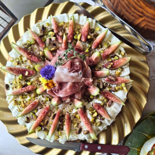 Guirlanda gastronômica da Alecrim disposta radialmente em uma bandeja dourada, composta por fatias de figos frescos, pistaches e flores comestíveis sobre base cremosa. No centro, o presunto cru dobrado de forma artística finaliza a iguaria, ideal para mesas gastronômicas e eventos de luxo em Belo Horizonte.