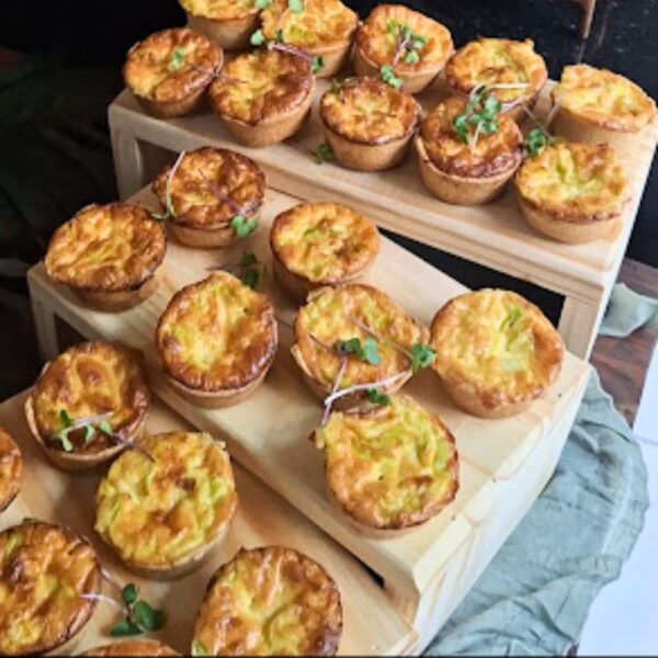 Mini quiches de alho-poró da Alecrim com recheio dourado e massa artesanal, decoradas com brotos verdes. Os finger foods estão organizados em suportes de madeira para um serviço de buffet gourmet, ideal para eventos corporativos e celebrações intimistas em Belo Horizonte.