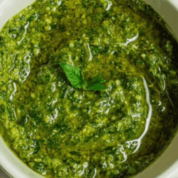 Molho pesto artesanal da Alecrim em close-up, com textura granulada de castanhas e queijo parmesão emersos em azeite de oliva vibrante. O molho é finalizado com uma pequena folha de manjericão fresco, representando a produção autoral e o cuidado com ingredientes selecionados para a gastronomia criativa em Belo Horizonte