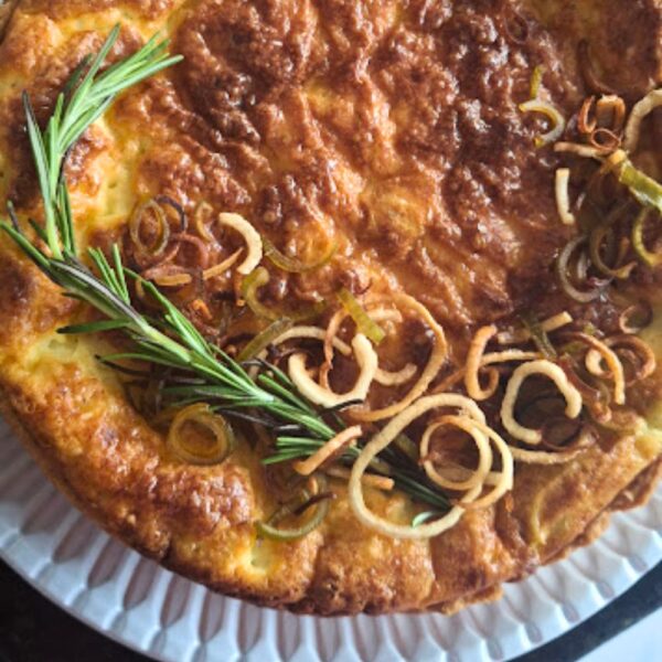 Quiche de alho-poró da Alecrim com cobertura dourada e crocante, decorada com anéis de alho-poró caramelizados e um ramo de alecrim fresco. O prato artesanal é servido em louça branca ornamentada, representando a gastronomia afetiva e sofisticada para eventos e recepções em Belo Horizonte.