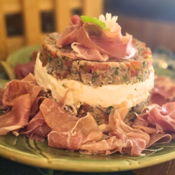 Torre de antepasto artesanal da Alecrim montada em camadas de cream cheese, tomates e ervas, decorada com fatias de presunto cru e flor comestível. Servida em prato de cerâmica verde, a iguaria exemplifica a gastronomia criativa e sofisticada para eventos e recepções em Belo Horizonte.