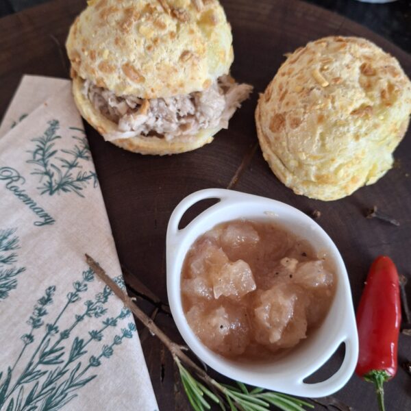 Sanduichinhos de pão de queijo da Alecrim recheados com pernil desfiado, acompanhados de chutney de abacaxi artesanal. Os mini sanduíches mineiros são apresentados em uma tábua rústica, ideal para coffee breaks, eventos e lanches gourmet em Belo Horizonte.
