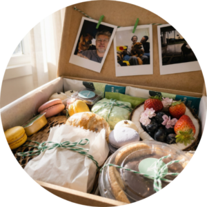 Cesta de café da manhã em BH servida em uma sofisticada caixa de madeira MDF com fotos polaroid personalizadas penduradas na tampa. O kit gourmet inclui macarons, pães artesanais, frutas frescas no coco e doces finos, unindo gastronomia autoral e memória afetiva para um presente único.