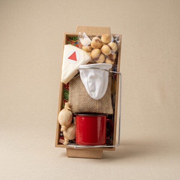 Caixa retangular em papel kraft, posicionada verticalmente em fundo neutro bege, contendo um kit gourmet organizado: xícara esmaltada vermelha, coador de pano com suporte metálico, pacote de biscoitos amanteigados, fatia de queijo, pequenos doces e itens artesanais, apresentados de forma limpa e centralizada com iluminação suave de estúdio.