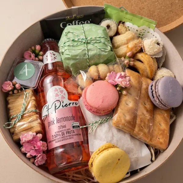 Caixa de presente de madeira redonda, vista de cima, preenchida com uma garrafa de Pink Lemonade da St. Pierre (275ml), um pacote de café da marca "Coffee", biscoitos artesanais amarrados com barbante, macarons nas cores rosa e lilás, pequenas flores cor-de-rosa e uma pequena etiqueta verde com o logotipo da letra