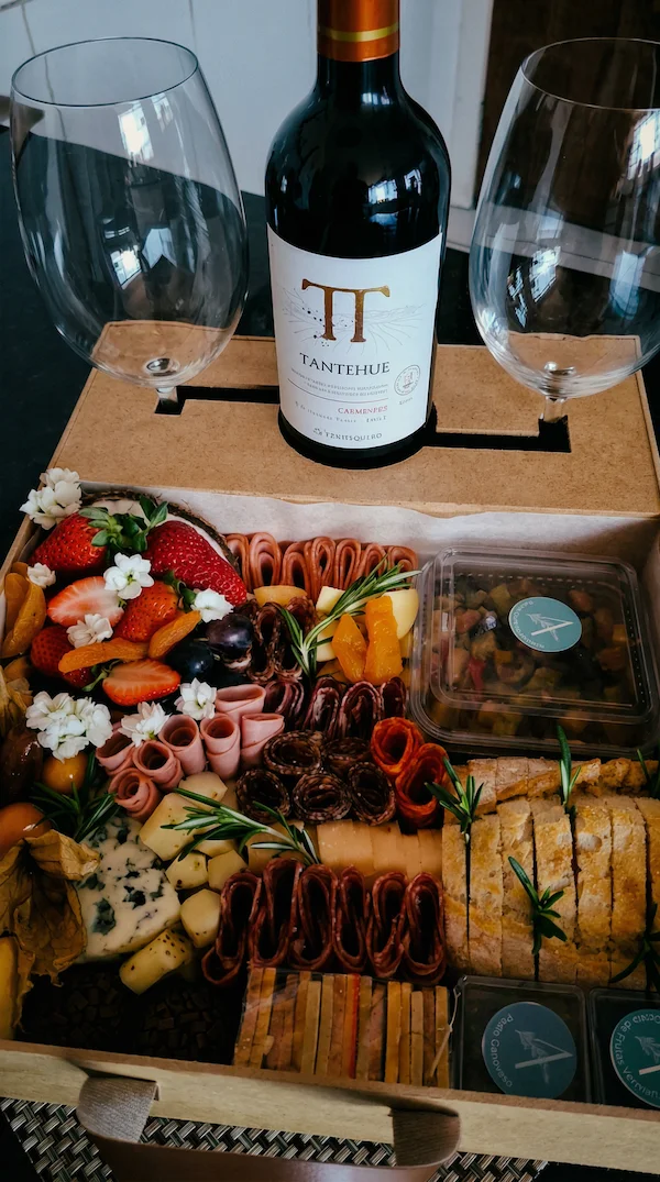 Experiência gourmet 'Quer Vin? Vem!' da Alecrim Gastronomia Criativa. Caixa de madeira artesanal com vinho importado, duas taças de vidro e tábua de charcutaria completa com queijos, salames, pães, antepastos e frutas frescas como morangos e physalis, decorada com alecrim.