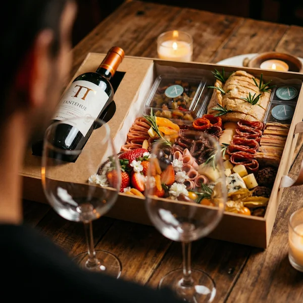 Experiência gourmet 'Quer Vin? Vem!' da Alecrim Gastronomia Criativa. Caixa de madeira artesanal com vinho importado, duas taças de vidro e tábua de charcutaria completa com queijos, salames, pães, antepastos e frutas frescas como morangos e physalis, decorada com alecrim.