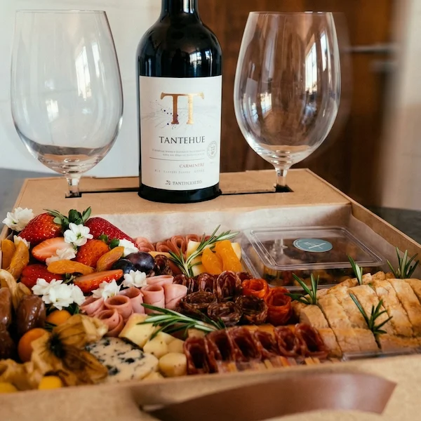 Experiência gourmet 'Quer Vin? Vem!' da Alecrim Gastronomia Criativa. Caixa de madeira artesanal com vinho importado, duas taças de vidro e tábua de charcutaria completa com queijos, salames, pães, antepastos e frutas frescas como morangos e physalis, decorada com alecrim.