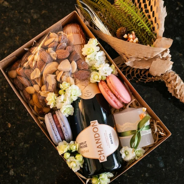 Detalhe dos macarons gourmet coloridos, mini buquê desidratado e vela perfumada com laço verde em um kit de presente afetivo e individual.