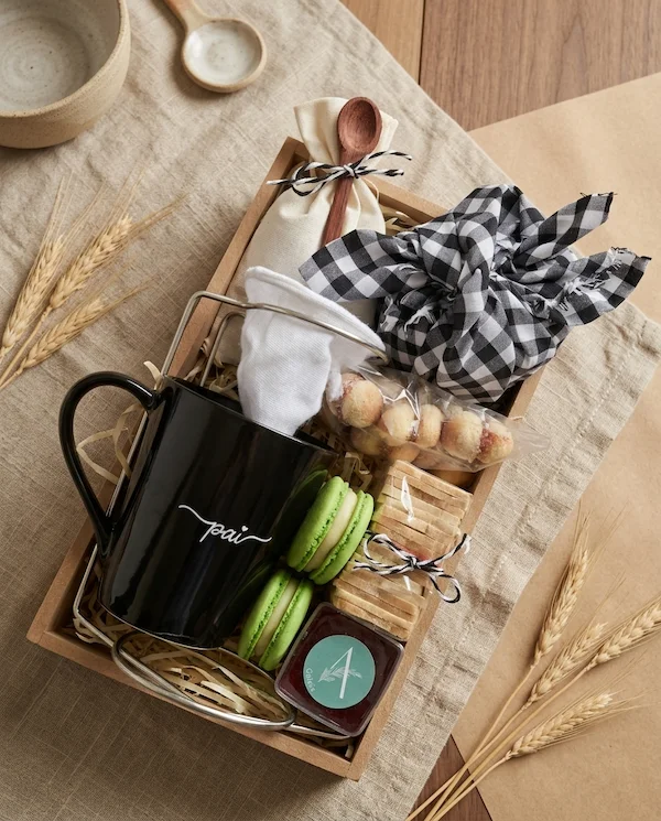 Cesta de café da manhã individual para o Dia dos Pais em BH da Alecrim Gastronomia Criativa, montada em caixa de madeira reutilizável com caneca preta personalizada 'Pai', coador de pano, macarons verdes, biscoitos artesanais e geleia caseira, decorada com trigo seco e tecido xadrez.