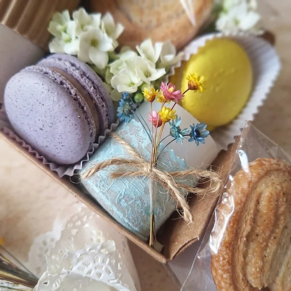 Detalhe aproximado de macarons de lavanda e limão, mini buquê de flores secas e palitos de folhado em uma caixa de presente gourmet Alecrim, destacando o cuidado artesanal e a estética afetiva.