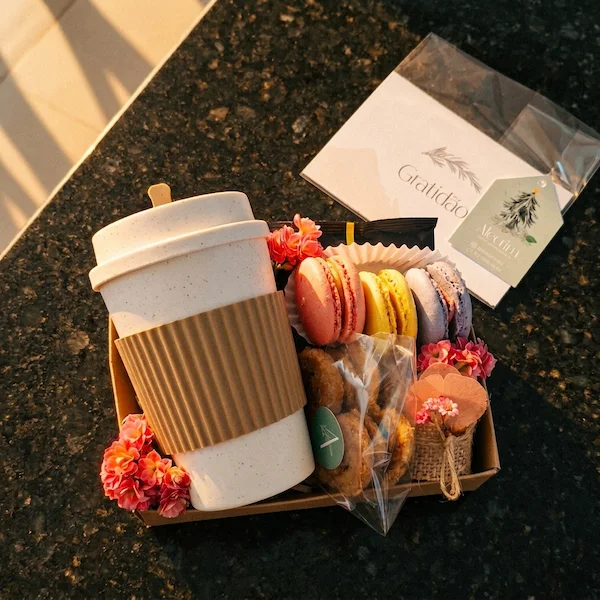 presente com café e doces da Alecrim Gastronomia Criativa em BH, contendo copo térmico branco com luva marrom, macarons coloridos, biscoitos artesanais e tag de agradecimento 'Gratidão', sobre bancada de granito com luz solar natural.